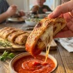 A close-up of a hand dipping a cheesy slice of stromboli into a bowl of marinara sauce with a cheese pull.