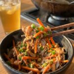 A close-up of chopsticks lifting a bite of pork and cabbage stir fry from a bowl.
