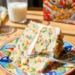 A close-up of a slice of cake showing rainbow cereal specks baked inside the sponge layers.