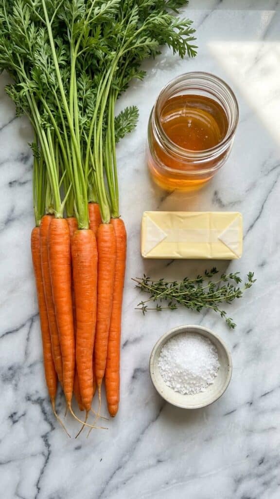 Overhead flat lay of ingredients: fresh carrots with tops, honey, butter, thyme, and sea salt on marble.