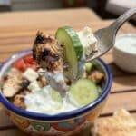 A close-up of a fork lifting a bite of chicken, feta, and tzatziki from a Greek bowl.