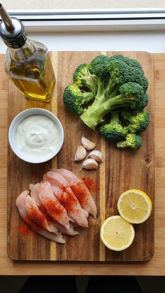 A flat lay showing seasoned raw chicken, fresh broccoli, greek yogurt, lemon, and garlic on a wooden board.