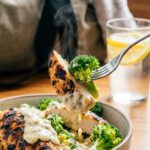 A close-up of a fork lifting grilled chicken and broccoli covered in white garlic sauce.