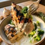 A close-up of a fork lifting grilled chicken and broccoli covered in creamy sauce.