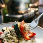 A close-up of a fork lifting grilled vegetables and cheese with a blurred outdoor BBQ background.
