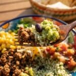 A close-up of a fork lifting a bite of beef, rice, and guacamole from a burrito bowl.