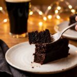 A close-up of a slice of dark chocolate stout cake with a fork, with a beer glass in the background.