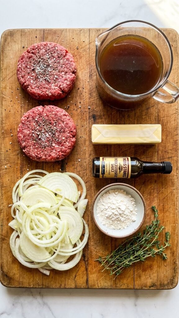Overhead flat lay view of ingredients: raw beef patties, sliced onions, beef broth, butter, flour, and Worcestershire sauce on a wooden board.