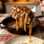 A close-up POV shot of a fork lifting a bite of hamburger steak dripping with rich onion gravy.
