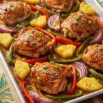 A sheet pan filled with roasted Hawaiian chicken thighs, caramelized pineapple chunks, and colorful bell peppers coated in a sticky glaze, garnished with sesame seeds.