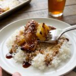 A close-up POV shot of a fork lifting a bite of glazed chicken and roasted pineapple over a bed of white rice.