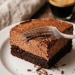 A close-up of a fork cutting into a very moist slice of dark chocolate cake with a glass of milk in the background.