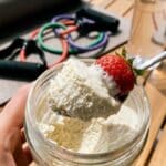 A close-up of a hand holding a cheesecake jar with a spoon lifting a creamy bite with fruit.