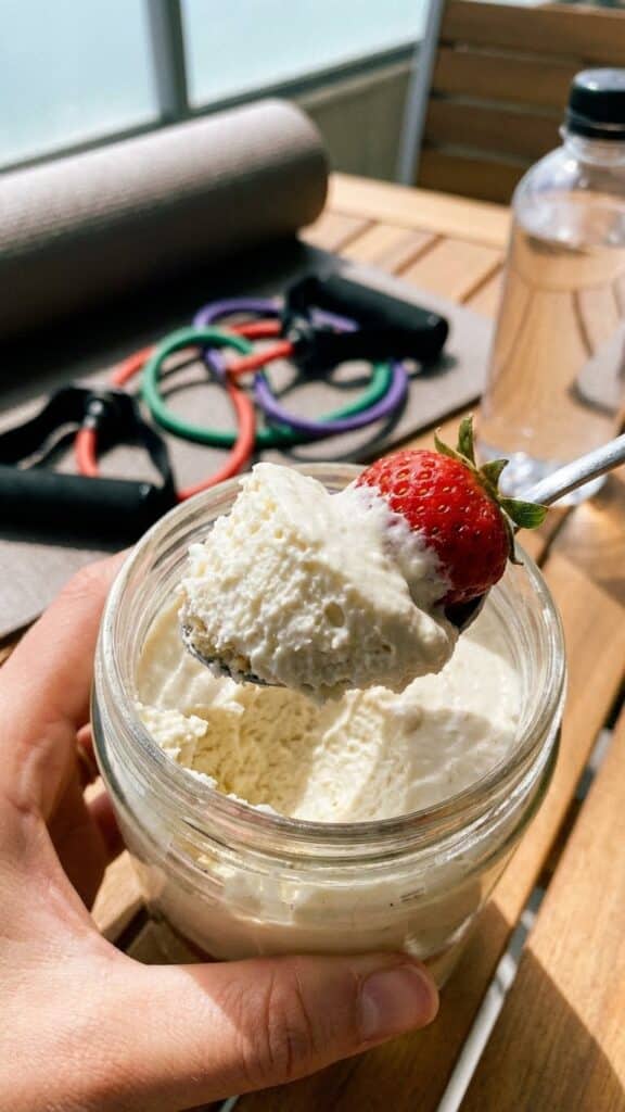 A close-up of a hand holding a cheesecake jar with a spoon lifting a creamy bite with fruit.