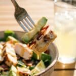 : A close-up of a fork lifting chicken, avocado, and bacon with dressing from a salad bowl.