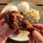 A close-up POV shot of hands holding a bitten Huli Huli chicken drumstick, showing juicy meat and sticky glaze.
