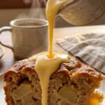 A close-up of vanilla custard being poured over a slice of apple cake, showing apple chunks inside.
