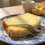 A flat lay of baking ingredients including a bowl of ricotta, fresh lemons with leaves, eggs, and flour on a stone surface.
