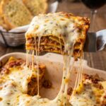 A spatula lifting a serving of ravioli lasagna with a long stretch of melted cheese