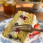 A close-up of a fork cutting into a slice of lemon cake with cream and berries.