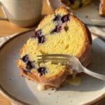 A close-up of a slice of lemon blueberry cake on a plate, showing the moist crumb and baked berries inside.