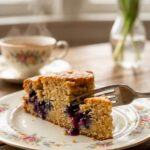 A close-up of a fork cutting into a slice of lemon blueberry cake on a floral plate.