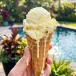 A hand holding a waffle cone with a scoop of melting lemon cottage cheese ice cream outdoors.