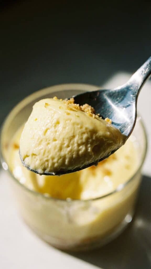 A close-up of a spoon lifting a scoop of thick, smooth lemon mousse with cracker crumbs.