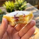 A close-up of a hand holding a lemon raspberry bar with a bite taken out, showing the gooey texture.