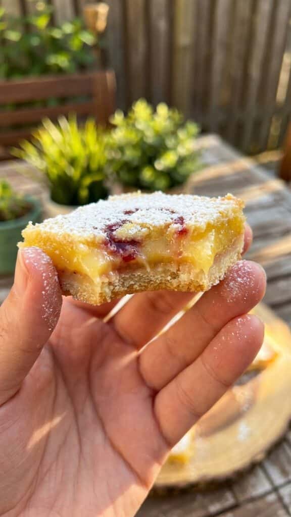 A close-up of a hand holding a lemon raspberry bar with a bite taken out, showing the gooey texture.
