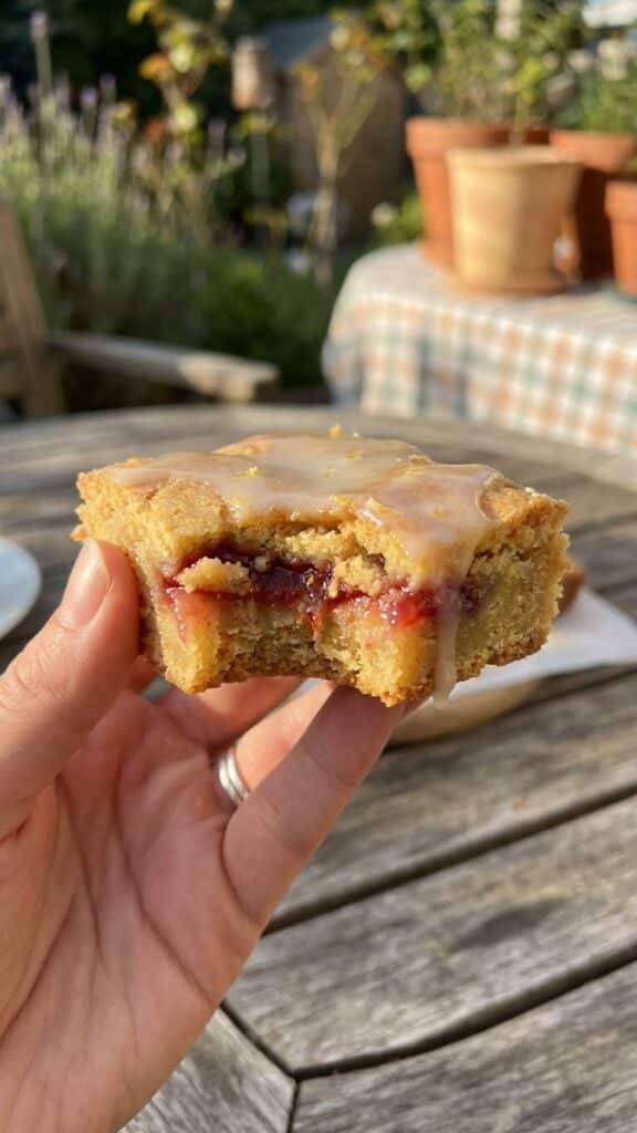 A close-up of a hand holding a strawberry lemon blondie with a bite taken out, showing the chewy texture and juicy fruit inside.