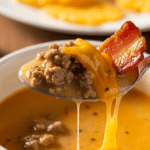 A close-up of a spoon lifting a bite of creamy beef and bacon soup from a bowl.
