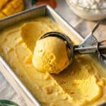 A close-up of an ice cream scoop curling a ball of mango cottage cheese ice cream from a pan.