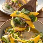 A close-up of a serving spoon lifting mango and cucumber slaw, with grilled food in the background.