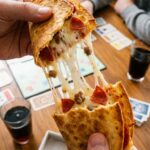 A close-up POV shot of hands pulling apart a pizza taco, revealing a long stretch of melted mozzarella cheese, pepperoni, and sausage.