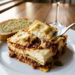 A close-up of a serving of ravioli casserole on a plate, showing the creamy filling and meat sauce layers with a fork.