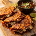 A close-up POV shot of a hand dipping a crispy beef and cheese taco into a bowl of red consommé broth.