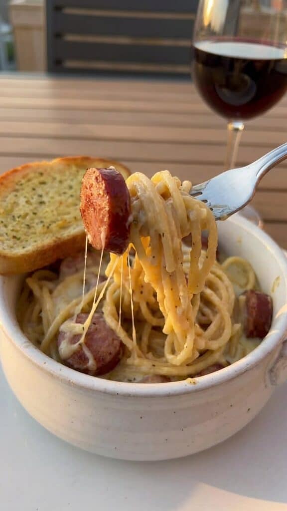 A close-up of a fork lifting a bite of creamy pasta and smoked sausage from a bowl.