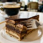 A close-up of a slice of opera cake with a fork cutting through the layers, served with coffee.