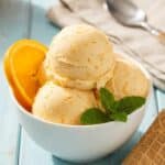A close-up of an ice cream scoop curling a ball of orange ice cream from a pan.