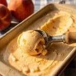 A close-up of an ice cream scoop curling a serving of peach ice cream from a loaf pan.