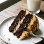 A close-up of a slice of chocolate cake with thick peanut butter frosting layers on a white plate.