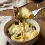 A close-up POV shot of a fork lifting a cheesy bite of pasta with steak and peppers, showing a gooey cheese pull.