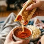 A close-up POV shot of a hand dipping a bitten pizza burrito into a bowl of marinara sauce.
