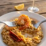 A close-up of a fork lifting a cooked prawn and creamy orzo pasta from a plate.