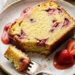 An overhead flat lay view of baking ingredients arranged on marble, including fresh strawberries in a colander, sticks of butter, sugar, eggs, flour, and a loaf pan.