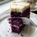 A close-up of a slice of purple cake with white frosting on a plate, with a fork taking a bite.