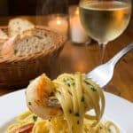 A close-up of a fork twirling linguine and a shrimp with a glass of wine in the background.