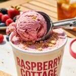A close-up of an ice cream scoop forming a ball of pink raspberry ice cream from a container.