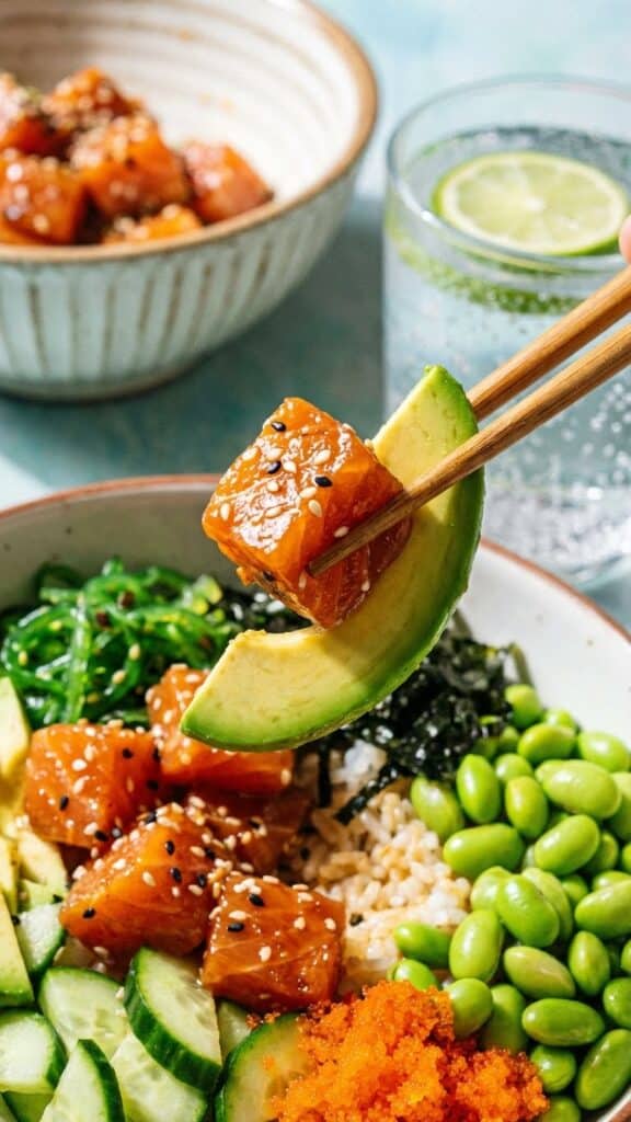 Close-up of chopsticks lifting a piece of marinated salmon and avocado from a poke bowl.